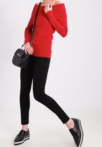 Maglione rosso off-the-shoulder, pantaloni neri aderenti, borsa a tracolla nera e scarpe nere con suola a strisce bianche. L'outfit è casual e moderno.
