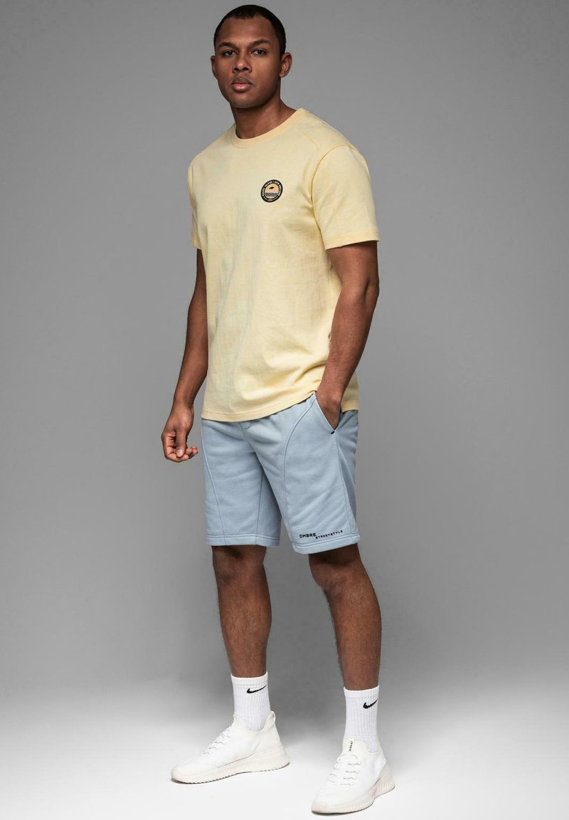 T-shirt en coton jaune avec logo rond, associé à un short bleu clair et des baskets blanches. Le mannequin pose les mains dans les poches sur un fond gris.