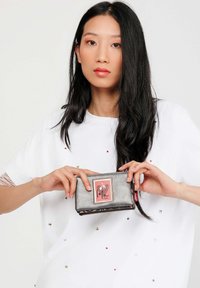 Portafoglio in pelle nera con etichetta grafica rosa, chiusura a zip e finitura testurizzata. Il modello lo tiene davanti a una camicia bianca con paillettes.