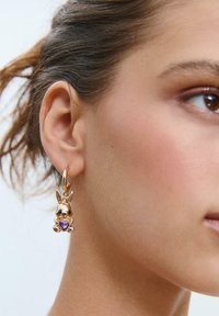Guldtonede metaløreringe med en kanincharme prydet med en lilla hjerteformet perle. Glat, skinnende overflade med detaljerede udsmykninger.