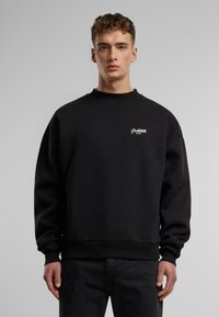 Czarny sweatshirt wykonany z mieszanki bawełny z ściągaczem w okolicy szyi i na mankietach. Posiada białe logo "Zabronione" na lewej stronie klatki piersiowej.
