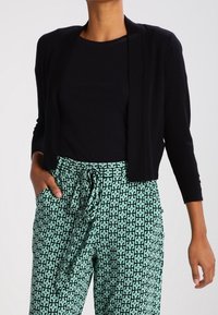 Cardigan noir ouvert sur le devant avec manches trois-quarts, associé à un pantalon vert à motifs géométriques avec un lien à la taille.