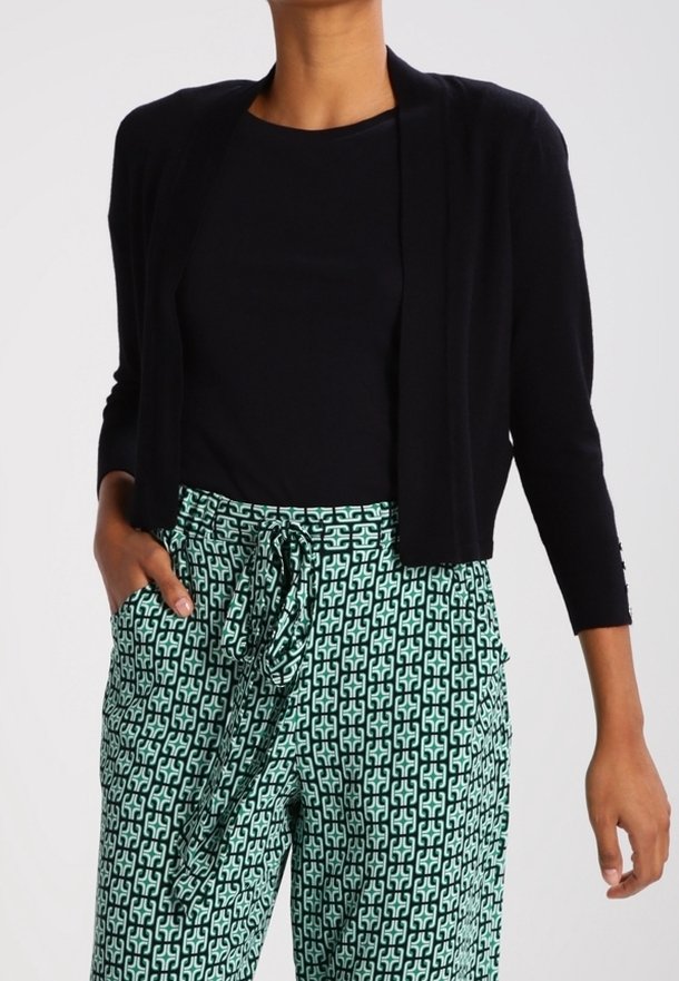 Cardigan noir ouvert sur le devant avec manches trois-quarts, associé à un pantalon vert à motifs géométriques avec un lien à la taille.