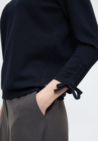 Granatowy sweter o fakturowanej strukturze i dopasowanym kroju, z ozdobnymi wiązaniami na rękawach oraz jednolitym granatowym paskiem na dole. Połączony z szarymi spodniami.