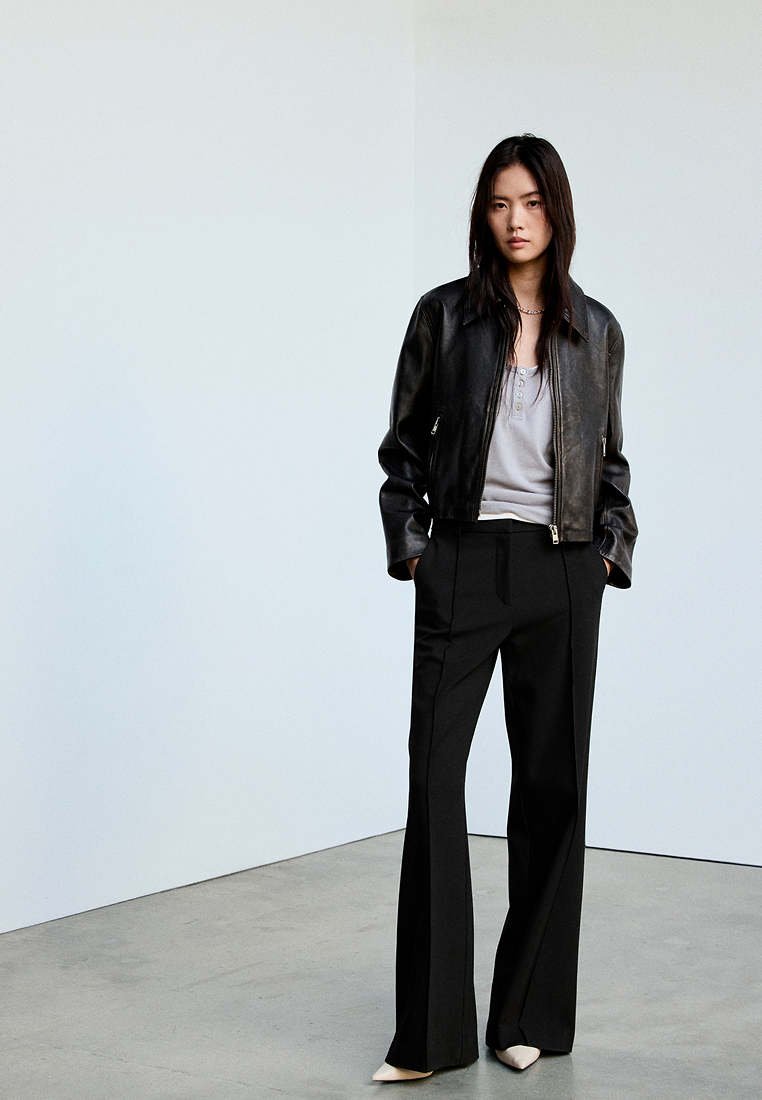 Femme portant une veste en cuir noire, un haut gris, un pantalon large noir et des chaussures blanches pointues, debout contre un mur clair uni.
