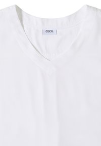 T-shirt blanc à col en V avec une petite étiquette "CECIL" cousue à l'intérieur du col, présenté à plat avec un focus sur l'encolure et la texture du tissu.