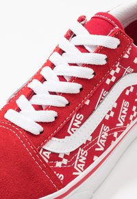 Baskets Vans rouges avec lacets blancs, bande latérale blanche et logo Vans répété sur un tissu à carreaux rouge et blanc.