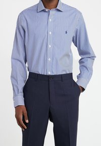 Camicia da uomo a maniche lunghe con strisce verticali blu e bianche, con bottone frontale e colletto classico. Indossata con pantaloni scuri.