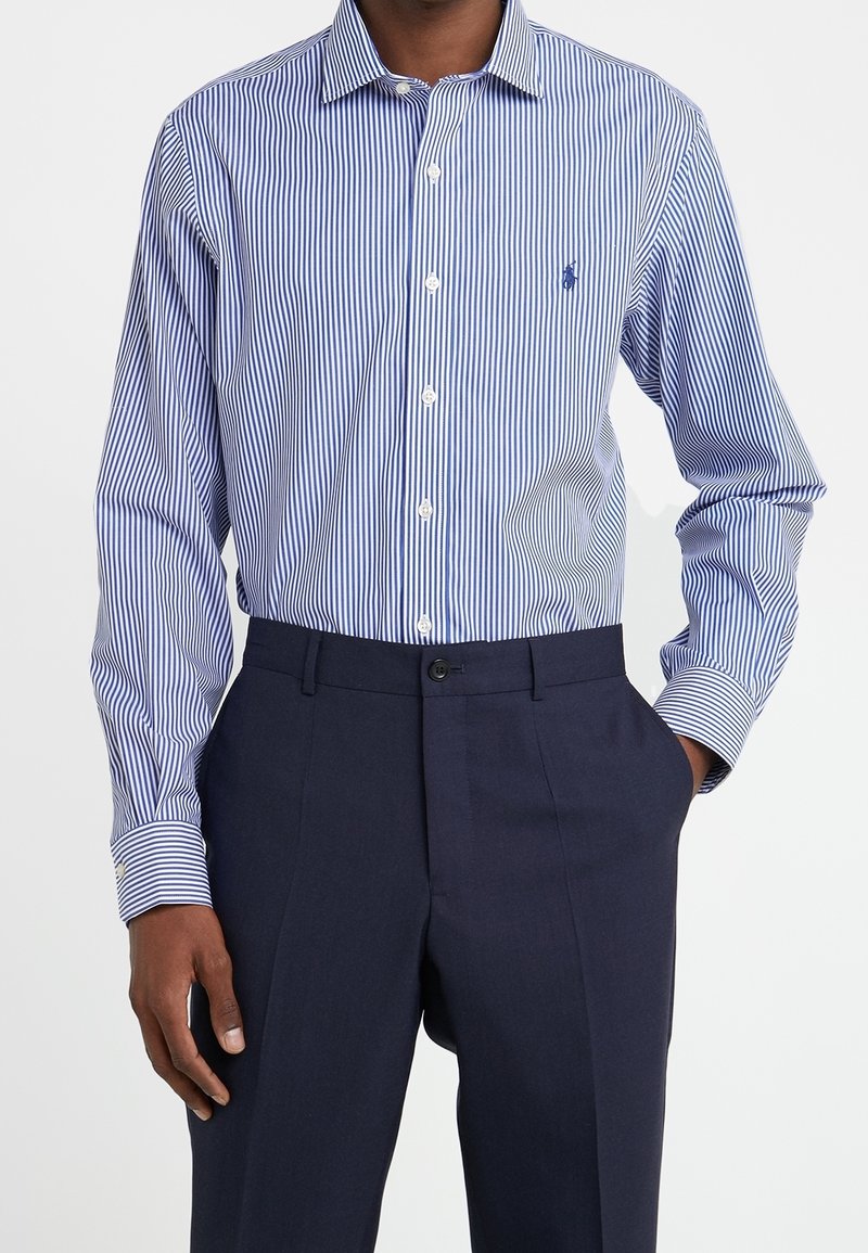 Camicia da uomo a maniche lunghe con strisce verticali blu e bianche, con bottone frontale e colletto classico. Indossata con pantaloni scuri.