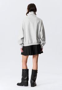 Szürke pulóver magas gallérral, laza szabással és bordázott szegéllyel. Fodros fekete szoknyával és térdig érő fekete csizmával párosítva.