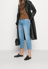 Gabardina negra de cuero sobre camisa marrón, vaqueros azules claros cortos y mocasines negros. Pequeño bolso negro con cadena como asa.