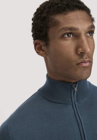 Blauer Strickpullover mit hohem Kragen und halbem Reißverschluss, der eine glatte Textur aufweist. Einfaches Design, das den Schwerpunkt auf Funktionalität legt.