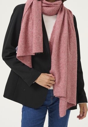 Personne portant une longue écharpe rose texturée sur un blazer noir, un haut blanc et un jean bleu, tenant l'écharpe d'une main.