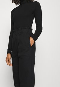 Černý rolák s dlouhými rukávy v kombinaci s volnými černými kalhotami s vysokým pasem, které mají kapsy a nařasený pas.