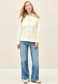 Valkoinen napitettava paita, jossa on kaksi rintataskua, wearattu harmaan paidan päällä, yhdistetty vaaleansinihin leveisiin farkkuihin ja valkoisiin lenkkareihin.