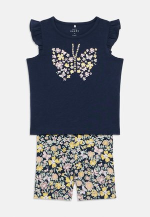 Canottiera in cotone blu navy con maniche arricciate e design floreale a farfalla; abbinata a pantaloni corti stampati con fiori nei toni del rosa e giallo su uno sfondo scuro.