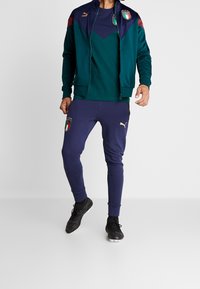 Puma trainingspak met een ritsjack in teal en maroon, gecombineerd met marineblauwe slim-fit broek. Inclusief Italiaans voetbal embleem en zwarte voetbalschoenen.