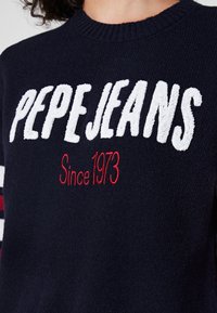 Pull en maille bleu marine avec l'inscription "PEPEJEANS" en lettres blanches en peluche et "Since 1973" en rouge, avec des accents rayés sur les manches.