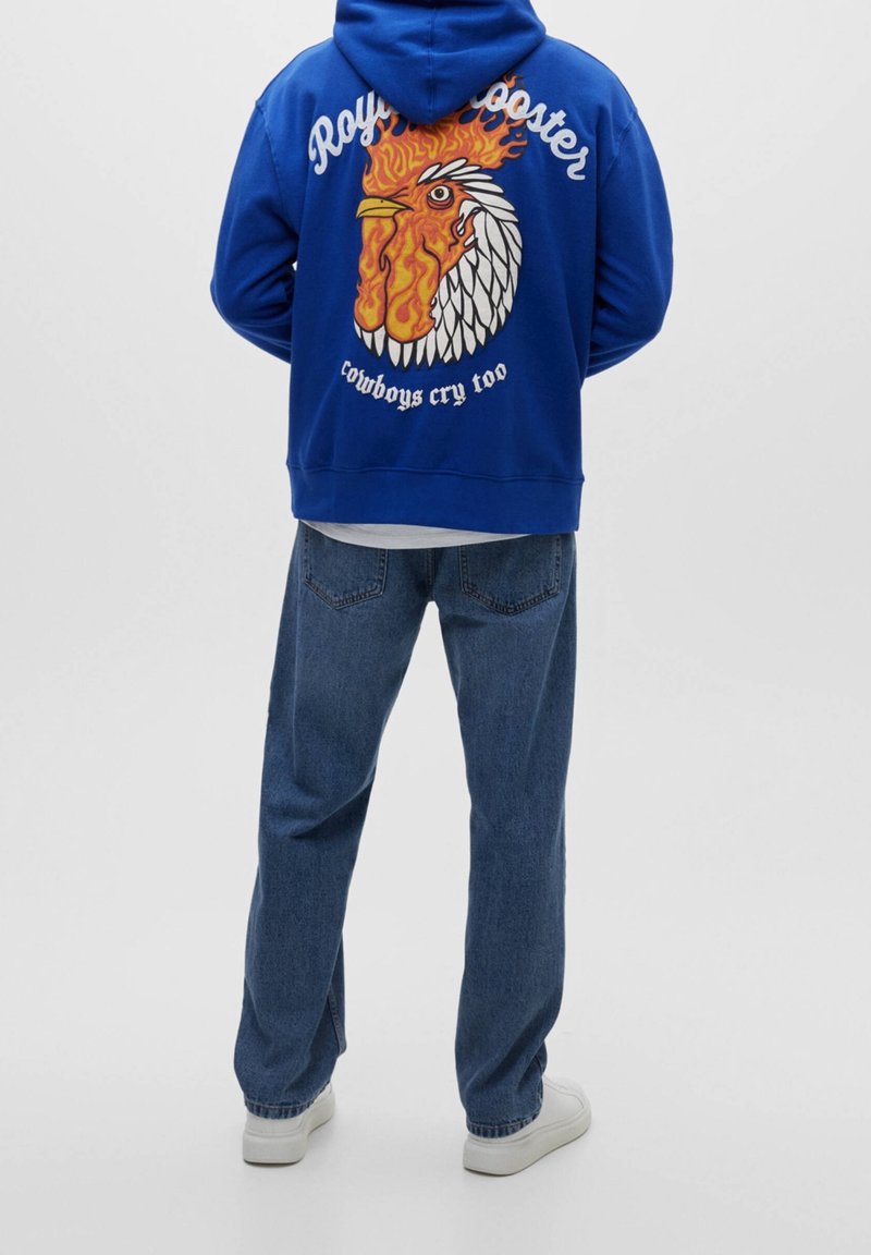 Blå hoodie med en grafisk bild av en tupp och lågor på ryggen, kombinerad med jeans och vita sneakers. Enkel design, avslappnad passform.