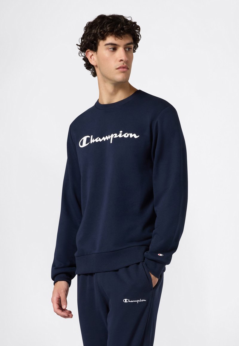 Sweatshirt bleu marine avec logo "Champion" blanc, col rond, manches longues, poignets et ourlet côtelés, fabriqué en tissu doux et texturé.