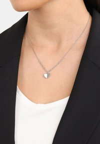 Collana con pendente a forma di cuore in argento, su una catena sottile, con finitura lucida e design a maglie alternate, indossata sopra una maglietta bianca e un blazer nero.