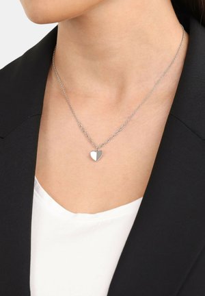 Collar de dije en forma de corazón de plata con una cadena delgada, que presenta un acabado pulido y un diseño de eslabones alternados, llevado sobre una blusa blanca y una chaqueta negra.