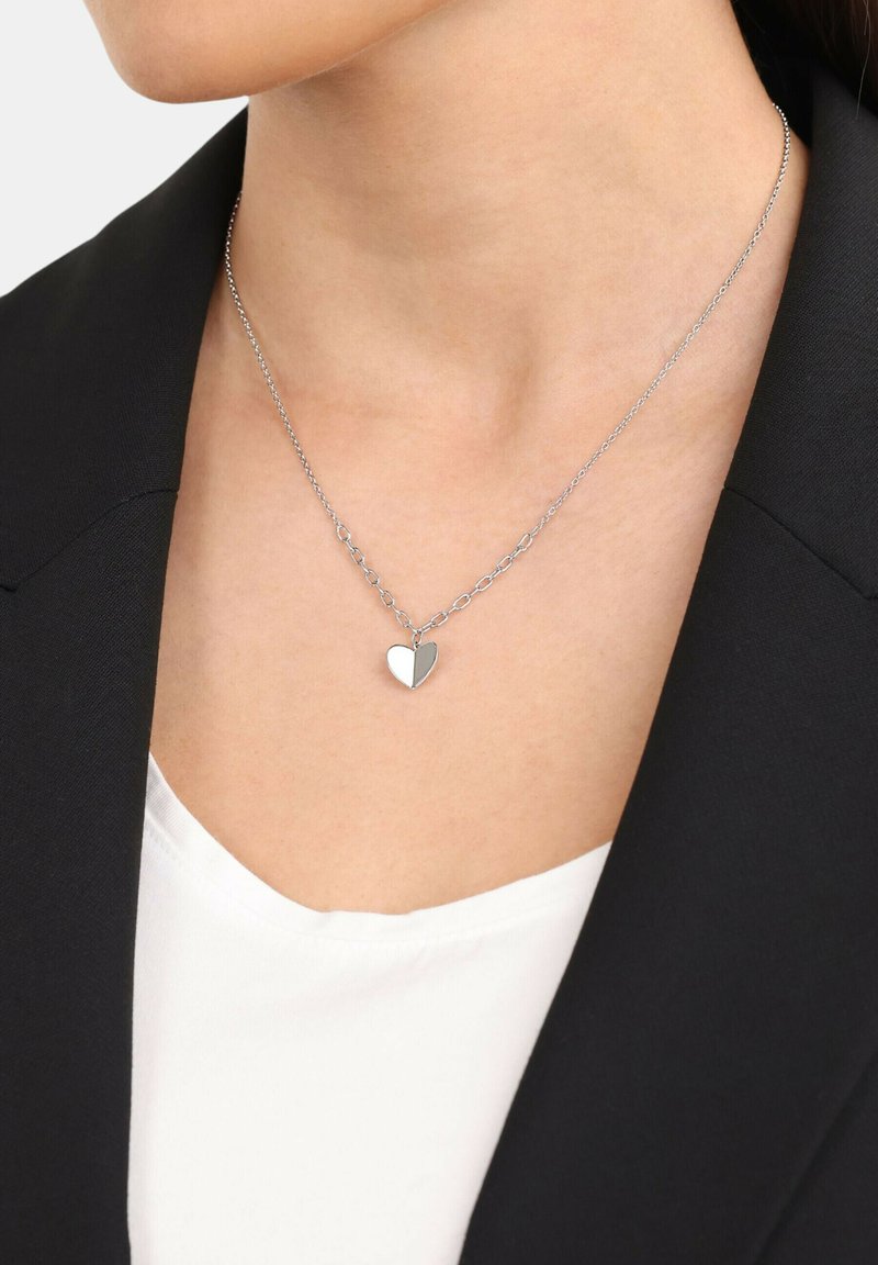 Collana con pendente a forma di cuore in argento, su una catena sottile, con finitura lucida e design a maglie alternate, indossata sopra una maglietta bianca e un blazer nero.