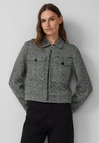 Kobieta z brązowymi włosami nosi czarno-białą, teksturowaną kurtkę na zamek z kołnierzem i czarne spodnie na szarym tle.