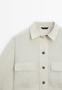 Light beige linen shirt with a button-up front, pointed collar, and two chest pockets. Features dark round buttons and a slightly textured fabric.