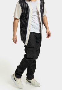 Camicia a righe bianche e nere con chiusura a bottoni, indossata sopra una canotta bianca. Pantaloni cargo neri e sneakers bianche con dettagli neri.