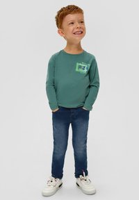 Groene langarmshirt met de opdruk "Always Fly Higher", gecombineerd met blauwe jeans en witte sneakers. Het materiaal lijkt zacht en casual.