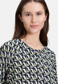 Granatowa bluzka z abstrakcyjnym wzorem w zielono-kremowe odcienie. Posiada okrągły dekolt i marszczenia przy ramionach dla luźniejszego kroju.