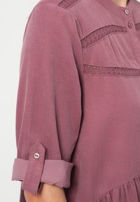 Blouse mauve à manches longues avec poignets roulés, patte à bouton, inserts en dentelle et détails plissés sur la poitrine et les épaules.