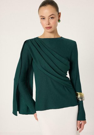 Vrouw draagt een donkergroene asymmetrische top met lange mouwen, geplooide stof en gespleten mouwen, gecombineerd met een witte rok en grote gouden en groene sieraden.