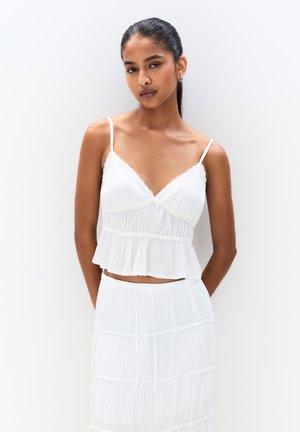Fehér, könnyű, redőzött cropped top vékony pántokkal és csipkés díszítéssel, ami egy illő, réteges szoknyával van párosítva. Egyszerű, puha textúra.