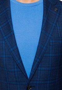 Granatowa marynarka w kratę o fakturowanym materiale, z niebieskimi akcentami i zapięciem na guzik, warstwowa na gładkiej niebieskiej koszulce z okrągłym dekoltem.