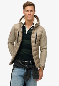 Beige puffjakke med hætte, som har sorte lynlåslommer og en khaki-farvet bluse med lange ærmer under. Båret med blå jeans.