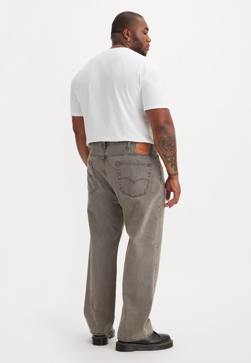Jeans larges gris avec un patch en cuir à l'arrière, portés avec un t-shirt blanc et des chaussures noires ; présentent des coutures visibles et des poches.