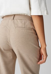 Beige broek met een gestructureerde afwerking, voorzien van een platte tailleband, zijzakken en dubbele achterzakken. De hand van het model rust op de heup.