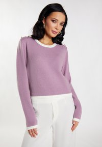 Lila trui met witte afwerking, lange mouwen en schouderknopen. Zachte textuur, cropped ontwerp, gecombineerd met witte broek.