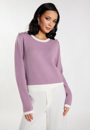 Lila Pullover mit weißen Akzenten, langen Ärmeln und Schulterknopfdetails. Weicher Stoff, cropped Design, kombiniert mit weißen Hosen.