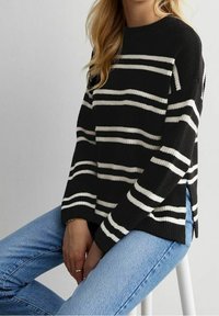 Frau in einem schwarzen Pullover mit weißen horizontalen Streifen, kombiniert mit hellblauen Jeans, sitzt auf einem weißen Hocker.