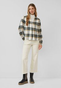 Camicia in flanella a quadri con un motivo verde, giallo e bianco, indossata sopra un dolcevita bianco, abbinata a pantaloni cropped color avorio e stivali neri.