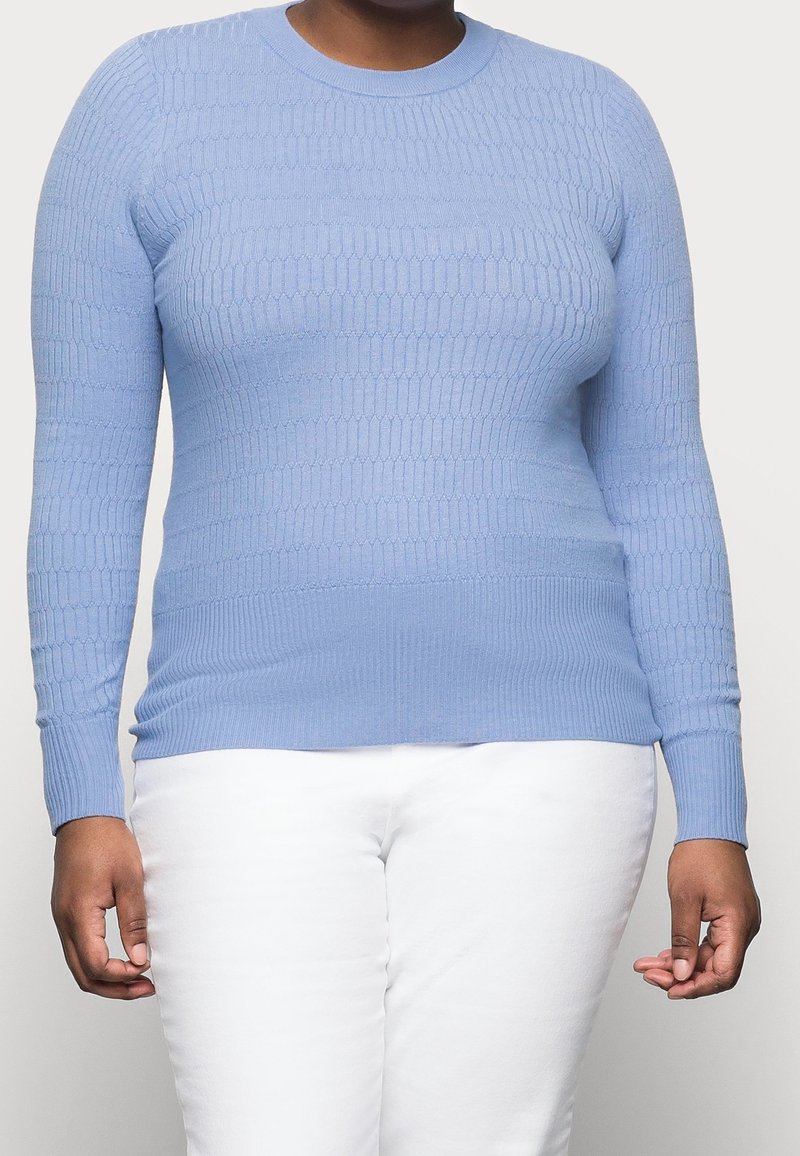 Hellblauer gestrickter Pullover mit Rippstruktur, langen Ärmeln und rundem Ausschnitt, kombiniert mit weißer Hose.