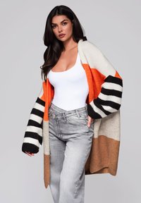 Sweter z dzianiny w pomarańczowe, czarne i białe paski, o luźnym kroju i teksturowanym materiale. Nosi się go na białym body i szarych dżinsach.