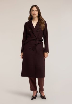 Mujer con un abrigo oscuro marrón con doble botonadura y cinturón, pantalones marrones con puños y tacones altos puntiagudos oscuros sobre un fondo liso.