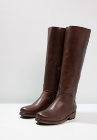 Bottes hautes en cuir marron avec une texture lisse, un bout arrondi et une fermeture éclair latérale. Talon en bois solide pour un soutien renforcé.