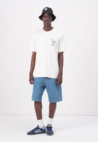 Hvit bomull T-skjorte med grafisk trykk på venstre bryst, kombinert med lyseblå denimshorts. Båret med svart bøttehatt og blå sneakers.