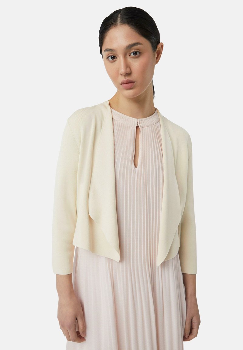 Crème-kleurige, cropped cardigan van zachte stof met een open voorkant, driekwartmouwen en subtiele textuur. Draagt over een geplooide roze jurk.