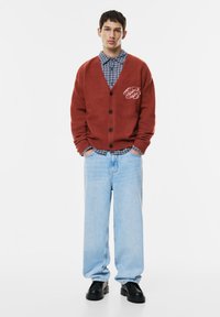 Rostfarbener Cardigan mit Knöpfen und Logo, über einem blau karierten Hemd getragen. Hellblaue weit geschnittene Jeans und schwarze Schuhe runden das Outfit ab.